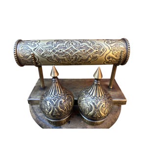 May include: A brass jewelry display stand featuring a horizontal cylindrical bar with intricate embossed patterns, supported by two vertical posts attached to a rectangular base, accompanied by two matching dome-shaped brass finials.