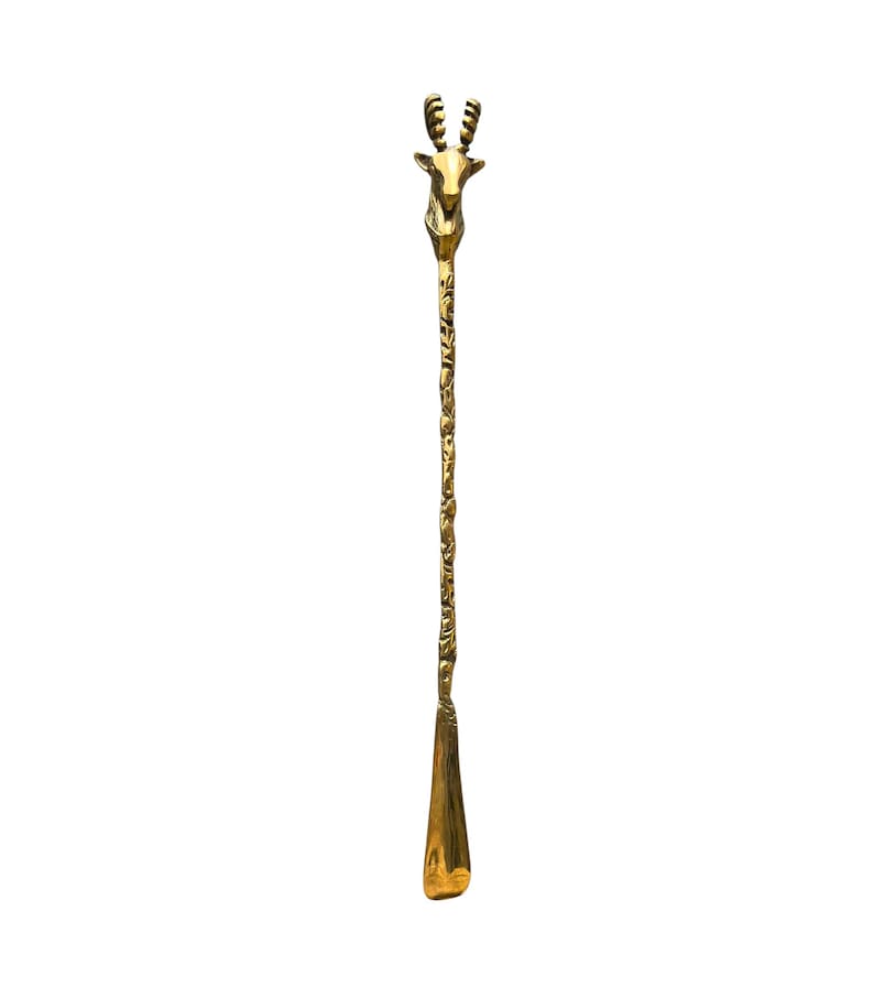 May include: A long, slender gold-toned metal shoehorn featuring a detailed deer head handle at the top. The handle transitions into a textured, patterned shaft that leads down to a smooth, curved scoop at the base for assisting with putting on shoes.