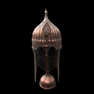 May include: A copper-colored, antique helmet with a domed top and a pointed finial. The helmet features decorative metalwork and a chainmail neck guard. The base has a bulbous shape, and the background is black.