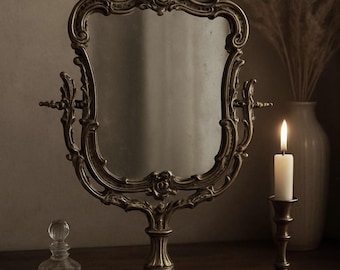 Puede incluir: Un espejo de tocador antiguo de latón con un marco ornamentado, un pequeño frasco de perfume de vidrio y una vela encendida en un candelabro de latón. Los objetos están dispuestos sobre una superficie de madera oscura, creando una estética vintage.