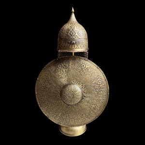 May include: An antique, ornate shield and helmet set. The shield is round with a central boss and intricate engraved patterns. The helmet is domed with a pointed top, also featuring detailed engravings. The set is made of a gold-colored metal.
