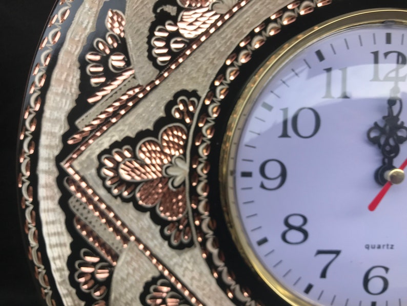 May include: A round, black and white clock with copper accents. The clock face is white with black numbers and hands. The clock is surrounded by a decorative border with a copper and white design. The clock says "quartz" at the bottom.