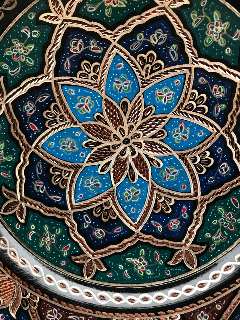 Puede incluir: Un plato decorativo con un patr&oacute;n geom&eacute;trico azul, verde y dorado. El plato tiene un fondo negro y un borde blanco.