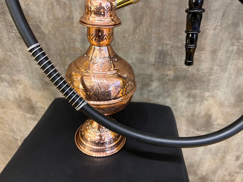 Op de afbeelding: Een koperen waterpijp met ingewikkelde ge&euml;tste ontwerpen. De waterpijp heeft een bolvormig lichaam, een decoratieve bovenkant en een zwarte slang met een zilveren spiraal. Het is een traditioneel rooktoestel.