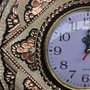 May include: A white clock face with black Roman numeral hour markers and black hands. The clock is set to 12:00. The clock is surrounded by a decorative copper and black metal frame.