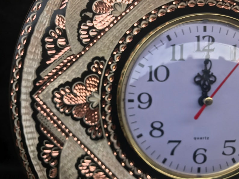 May include: A white clock face with black Roman numeral hour markers and black hands. The clock is set to 12:00. The clock is surrounded by a decorative copper and black metal frame.