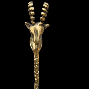 May include: A polished brass spoon featuring a handle shaped like the head and neck of an antelope. The animal head is detailed with prominent, curved, ridged horns and pointed ears. The stem of the spoon has a textured, organic, bark-like finish.