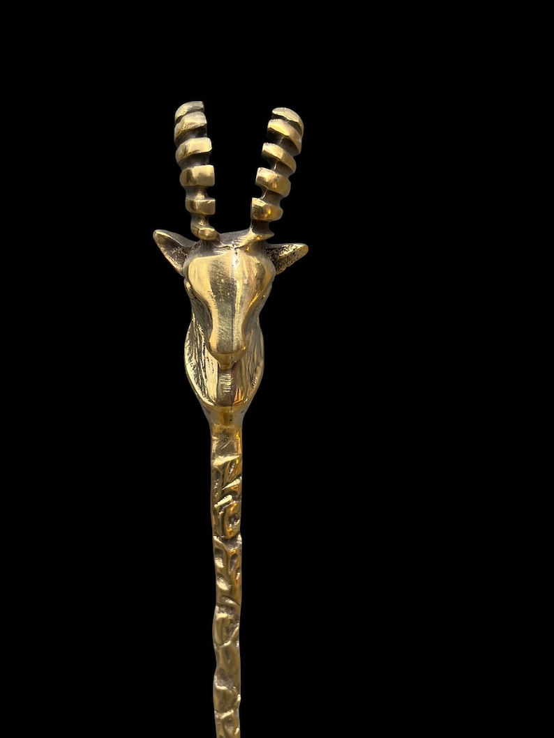 May include: A polished brass spoon featuring a handle shaped like the head and neck of an antelope. The animal head is detailed with prominent, curved, ridged horns and pointed ears. The stem of the spoon has a textured, organic, bark-like finish.