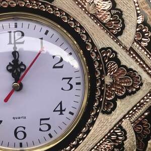 May include: A black and white clock with gold accents and a white face. The clock has Roman numerals and a red minute hand. The clock is surrounded by a decorative design with copper accents.