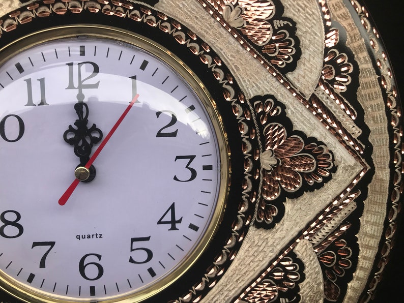 May include: A black and white clock with gold accents and a white face. The clock has Roman numerals and a red minute hand. The clock is surrounded by a decorative design with copper accents.