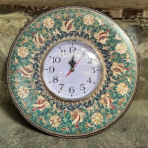 Può includere: Un orologio da parete rotondo con un quadrante bianco e numeri neri. L'orologio ha una cornice decorativa con un design floreale verde, oro e rosso.