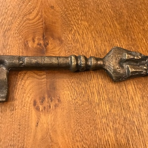 Könnte beinhalten: Ein dekorativer Schlüssel in Bronzefarbe mit einem verzierten Griff und einem detaillierten Bart. Der Schlüssel liegt auf einer Holzoberfläche. Der Schlüsselgriff hat ein einzigartiges, strukturiertes Design, und der Bart hat eine markante Form.