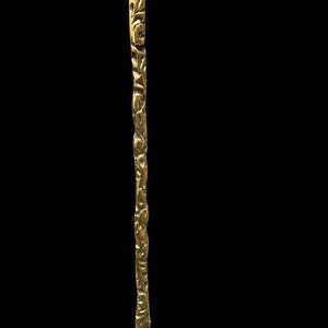 May include: A long, slender, metallic gold-toned stirring rod or swizzle stick featuring a textured, organic, branch-like surface pattern. The object has a polished finish that reflects light, with a slightly tapered end and a wider, rounded top section.