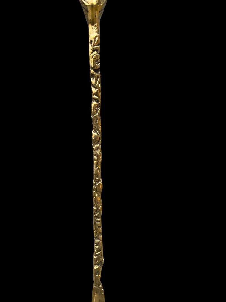 May include: A long, slender, metallic gold-toned stirring rod or swizzle stick featuring a textured, organic, branch-like surface pattern. The object has a polished finish that reflects light, with a slightly tapered end and a wider, rounded top section.