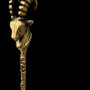 May include: A metallic gold-toned hair stick featuring a sculpted ibex head at the top. The ibex has prominent curved, ridged horns and detailed facial features. The shaft of the hair stick has a textured, uneven surface with a rustic finish.