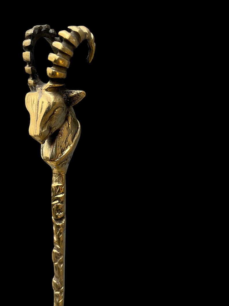 May include: A metallic gold-toned hair stick featuring a sculpted ibex head at the top. The ibex has prominent curved, ridged horns and detailed facial features. The shaft of the hair stick has a textured, uneven surface with a rustic finish.