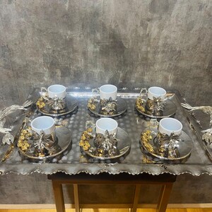 May include: A silver serving tray holding six teacups and saucers. Each saucer features a floral design with gold detailing. The white teacups have silver handles. The tray has decorative handles and a scalloped edge. A luxurious tea set.