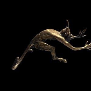 May include: A bronze sculpture of a demonic figure with horns, a long tail, and outstretched arms. The figure is in a dynamic pose, as if leaping or falling, against a black background. The sculpture has a detailed texture.