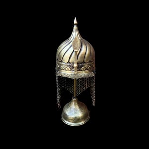 Op de afbeelding: Decoratieve messing helm met een gewelfde bovenkant, ingewikkelde details en een maliënkolder nekbescherming. De helm is gemonteerd op een bijpassende messing standaard. Zwarte achtergrond.