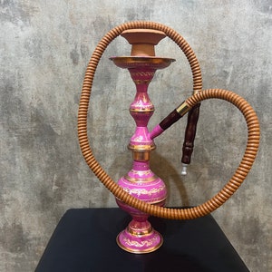 May include: A pink and gold hookah with a brown hose. The hookah has intricate designs and a round base.