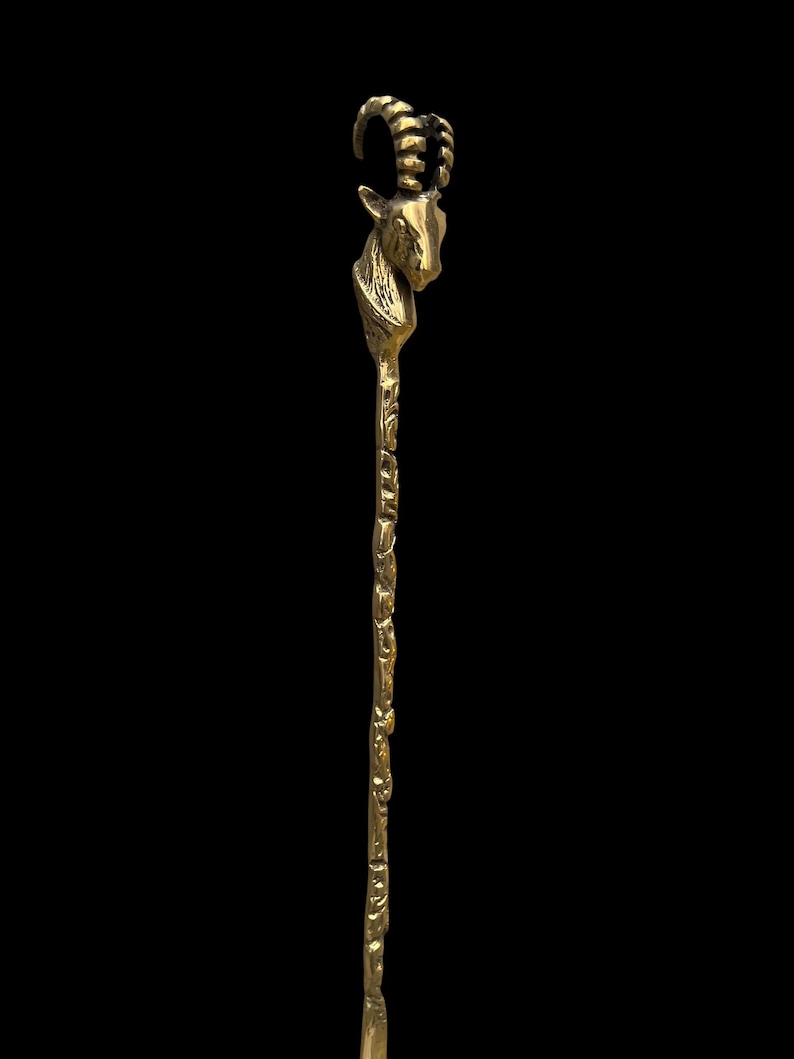 May include: A gold-toned metal hair stick featuring a sculpted goat head at the top with curved, ridged horns. The long, slender shaft of the hair accessory has a textured, uneven surface that mimics the appearance of a natural, organic branch.