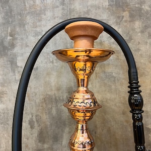 Op de afbeelding: Een koperen waterpijp met ingewikkelde ge&euml;tste ontwerpen en een terracotta kom. De waterpijp heeft een zwarte slang en een decoratieve steel. De afbeelding is tegen een getextureerde grijze achtergrond.