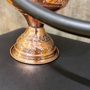 Op de afbeelding: Een koperkleurige decoratieve kom met een gedetailleerd ge&euml;tst ontwerp. De kom staat op een kleine basis met een decoratief patroon. Een donker metalen handvat buigt over de kom.