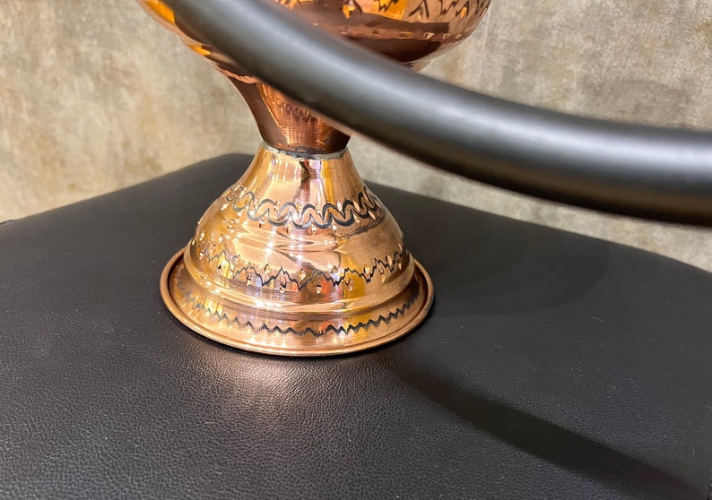 Op de afbeelding: Een koperkleurige decoratieve kom met een gedetailleerd ge&euml;tst ontwerp. De kom staat op een kleine basis met een decoratief patroon. Een donker metalen handvat buigt over de kom.