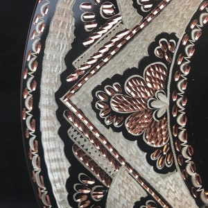 May include: A black and white circular plate with a copper-coloured design. The design features intricate floral patterns and geometric shapes.