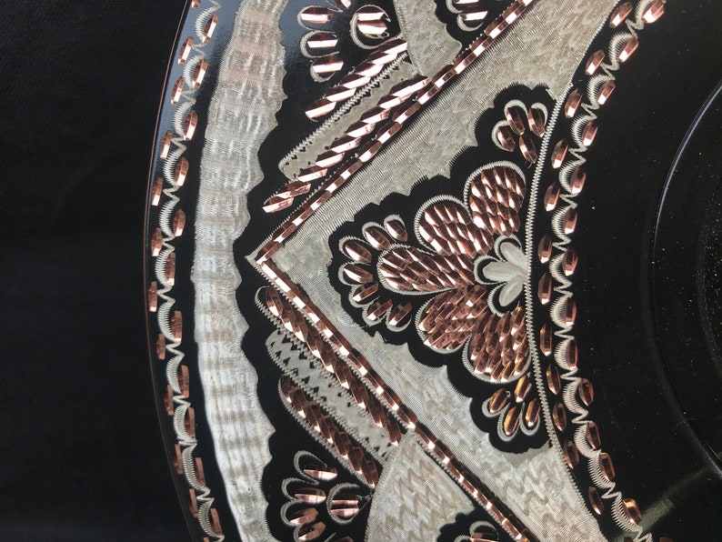 May include: A black and white circular plate with a copper-coloured design. The design features intricate floral patterns and geometric shapes.