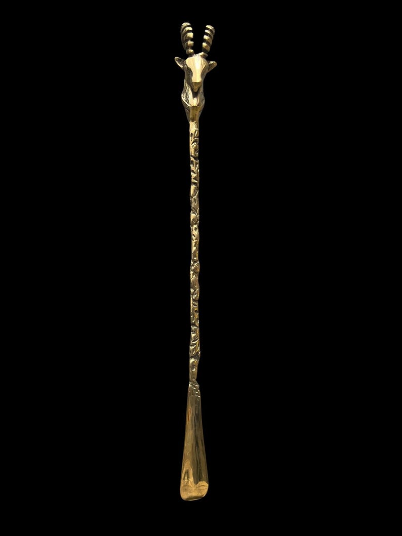 May include: A gold-colored metal shoehorn featuring a long, slender handle with an intricate, embossed floral pattern. The top of the handle is shaped like the head of an antelope with curved horns, while the bottom features a smooth, curved scoop.