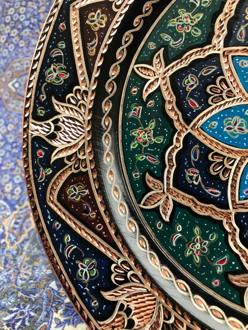 Puede incluir: Un plato decorativo redondo con intrincados dise&ntilde;os florales en tonos azules, verdes y rojos. El plato tiene un borde de color cobre con un patr&oacute;n detallado.