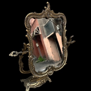 May include: Ornate antique vanity mirror with a decorative brass frame and stand. The mirror has a rectangular shape with a curved top and is supported by an elaborate base. The frame features intricate scrollwork and embellishments.