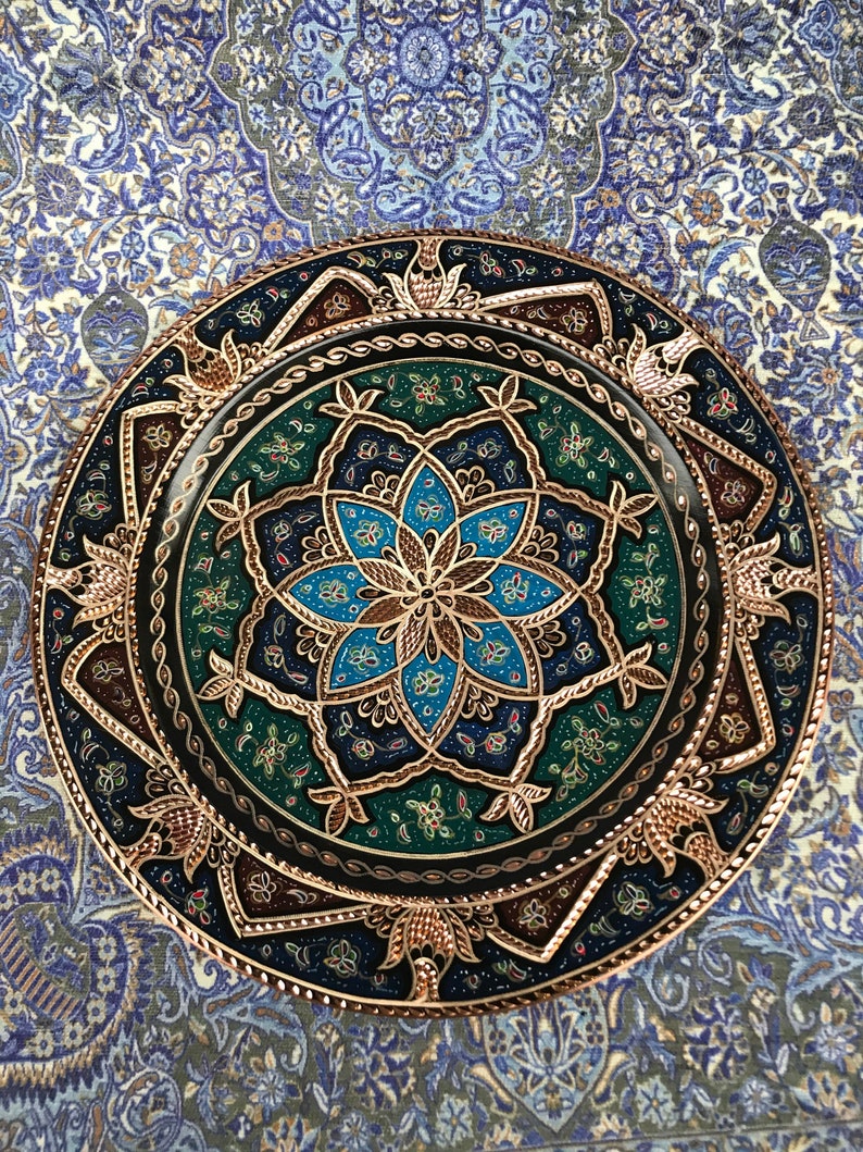 Puede incluir: Un plato decorativo redondo con fondo negro y dise&ntilde;os florales intrincados en oro, azul y verde. El plato tiene un borde de cobre y est&aacute; decorado con peque&ntilde;os detalles brillantes.