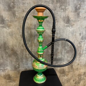 May include: An ornate green hookah with gold accents and a black hose. The hookah has a bulbous base and a slender, decorated body. The top features a brown bowl. The hookah is likely used for smoking flavored tobacco.