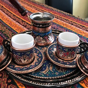 Op de afbeelding: Een traditionele Turkse koffieset met een koperen pot, twee kopjes en schoteltjes, allemaal versierd met ingewikkelde blauwe en gouden bloemmotieven. De set wordt gepresenteerd op een zwarte dienblad met een decoratieve rand.