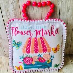 May include: A white pillow with a pink border and white pom-poms. The pillow has a floral design with a flower cart and butterflies. The text "always fresh Flower Market" is printed on the pillow.