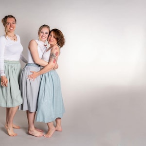 May include: Three people wearing white tops and skirts in shades of blue and gray. The skirts are knee-length and have a pleated design. The women are standing close together, smiling, and appear to be embracing. The background is a plain white.