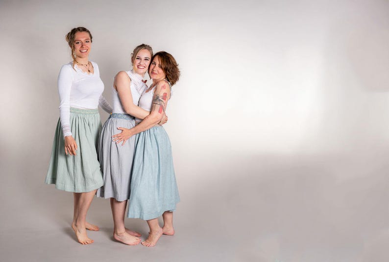 May include: Three people wearing white tops and skirts in shades of blue and gray. The skirts are knee-length and have a pleated design. The women are standing close together, smiling, and appear to be embracing. The background is a plain white.