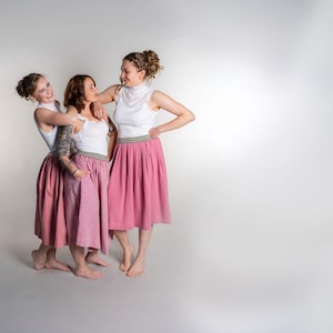 May include: Three people wearing matching outfits stand together. Each person wears a white top with a high neckline and a pink skirt. The skirts have a decorative band at the waist. The background is a solid light gray.