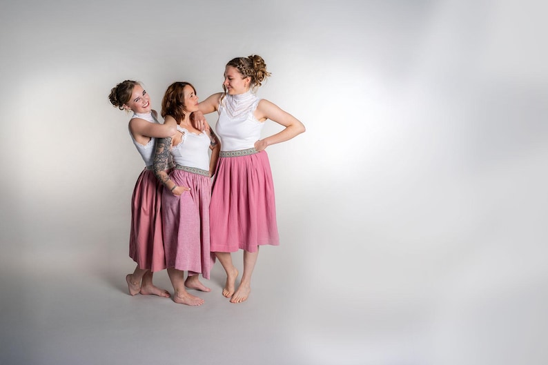 May include: Three people wearing matching outfits stand together. Each person wears a white top with a high neckline and a pink skirt. The skirts have a decorative band at the waist. The background is a solid light gray.