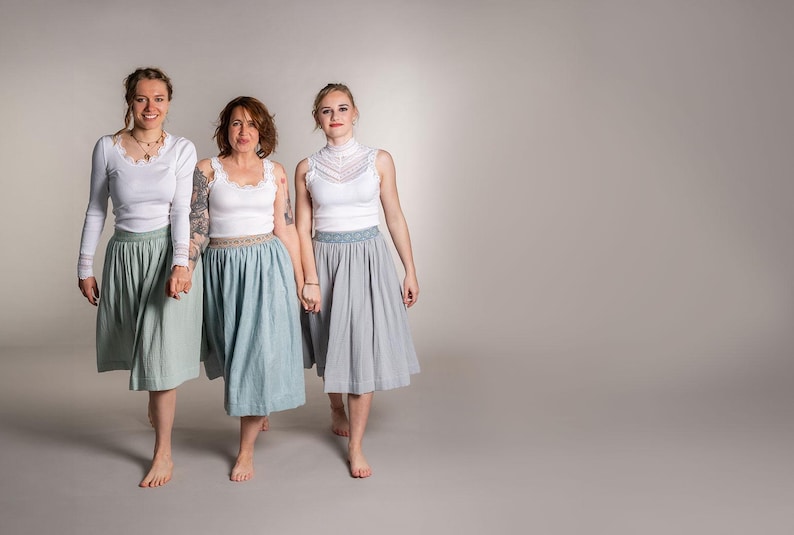 May include: Three people wearing white tops and skirts in shades of green, blue, and gray. The skirts are knee-length and have a gathered design. The tops include long-sleeved, tank, and high-neck styles. The women are holding hands and walking forward.