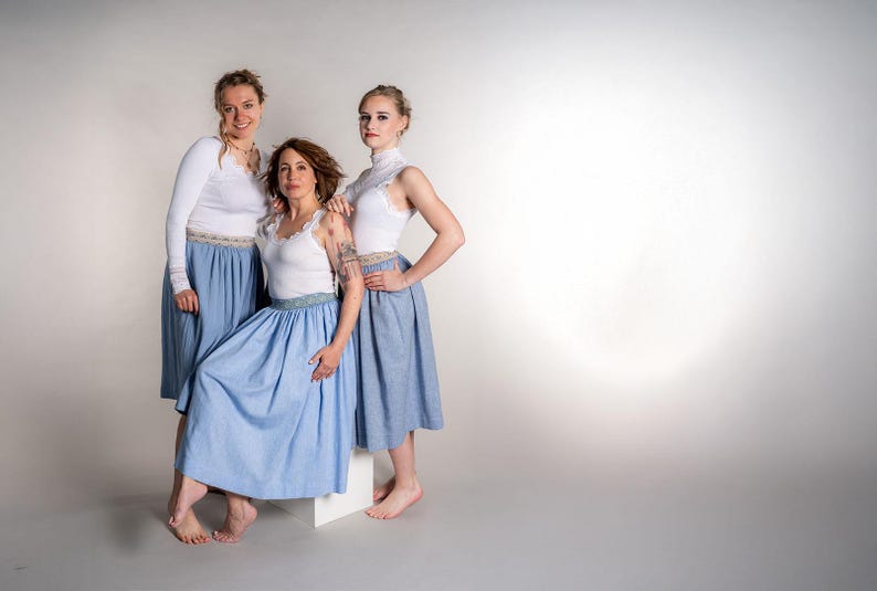 May include: Three people wearing matching outfits. Each person is wearing a white top and a light blue skirt. The skirts have a decorative band at the waist. The outfits are suitable for a dance or performance.