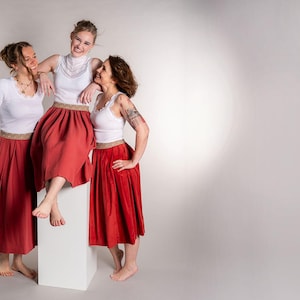 May include: Three people wearing white tops and red skirts pose against a white backdrop. The skirts are a vibrant red with a gold trim at the waist. One person is standing on a white block, while the others stand beside them.