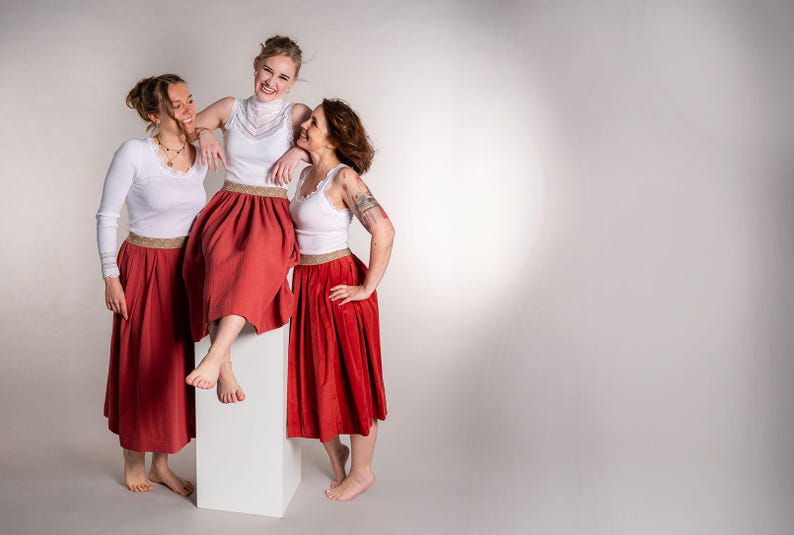 May include: Three people wearing white tops and red skirts pose against a white backdrop. The skirts are a vibrant red with a gold trim at the waist. One person is standing on a white block, while the others stand beside them.