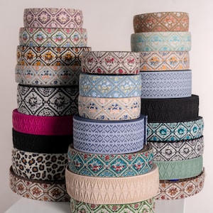Könnte beinhalten: Eine Sammlung dekorativer Bänder in verschiedenen Farben und Mustern. Die Bänder sind in Rollen gestapelt und zeigen florale, geometrische und Tiermuster. Farben sind Rosa, Schwarz, Beige und Türkis. Die Bänder eignen sich für Bastelarbeiten und Verzierungen, mit einer Höhe von ca. 5 cm pro Rolle.
