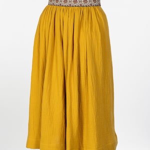 May include: Mustard yellow skirt with a patterned waistband. The skirt is made of a textured fabric and has a gathered waist. The waistband features a repeating geometric design in shades of brown, red, and cream.