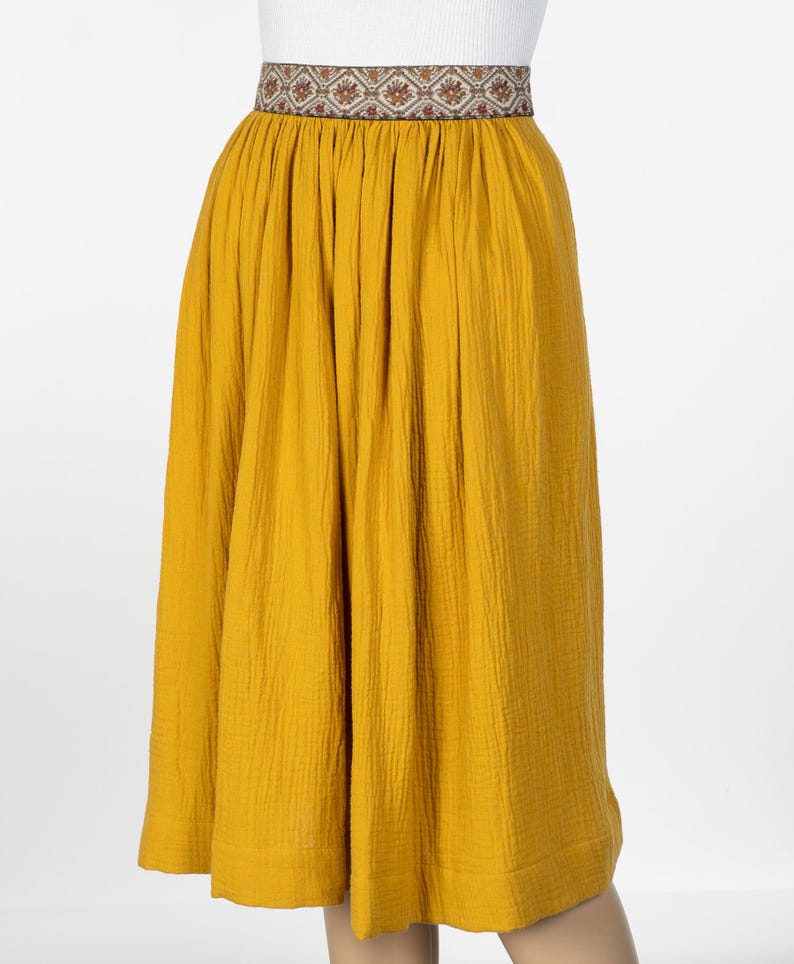 May include: Mustard yellow skirt with a patterned waistband. The skirt is made of a textured fabric and has a gathered waist. The waistband features a repeating geometric design in shades of brown, red, and cream.