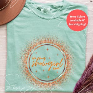 May include: A light green t-shirt with the text "in my showgirl era" in an orange and gold design. The shirt is folded on a white surface, with a brown hat and dried flowers in the background.