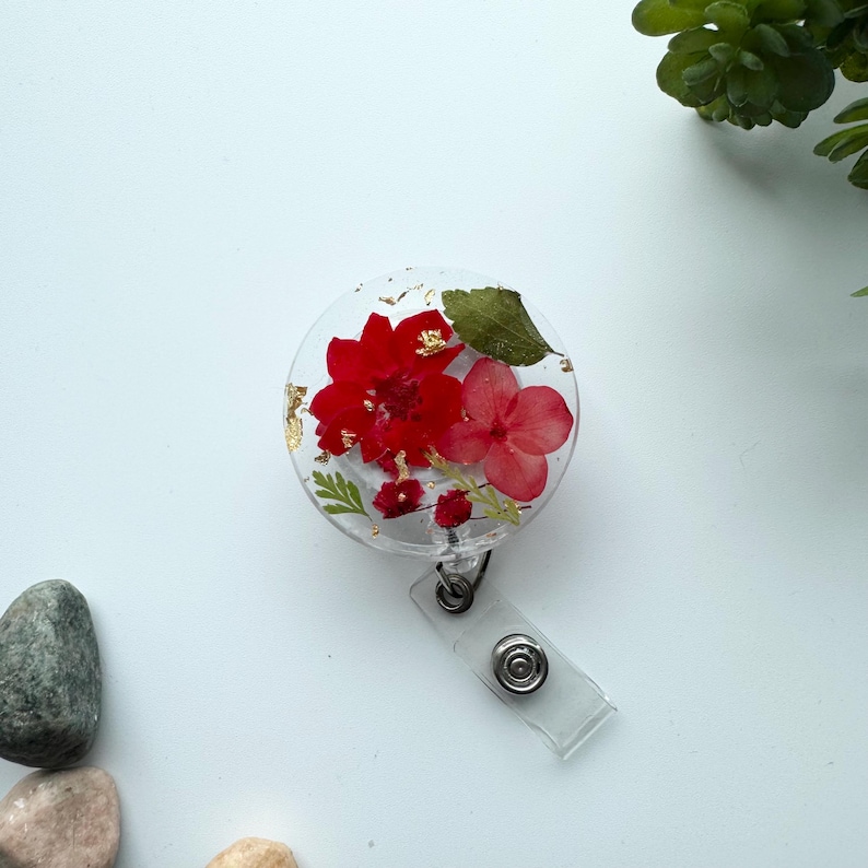 以下が含まれることがあります： 赤い花とピンクの花が押し花でデザインされた、透明な丸いバッジホルダー。金箔のアクセントが施されています。バッジホルダーは、リトラクタブルバッジリールに接続されています。